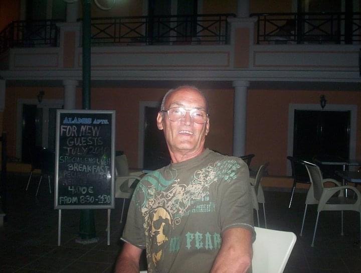 Picture of Just A Theatre Guys Father, Alan, sat down outside on holiday. Big smile on his face wearing a green shirt with skulls on.